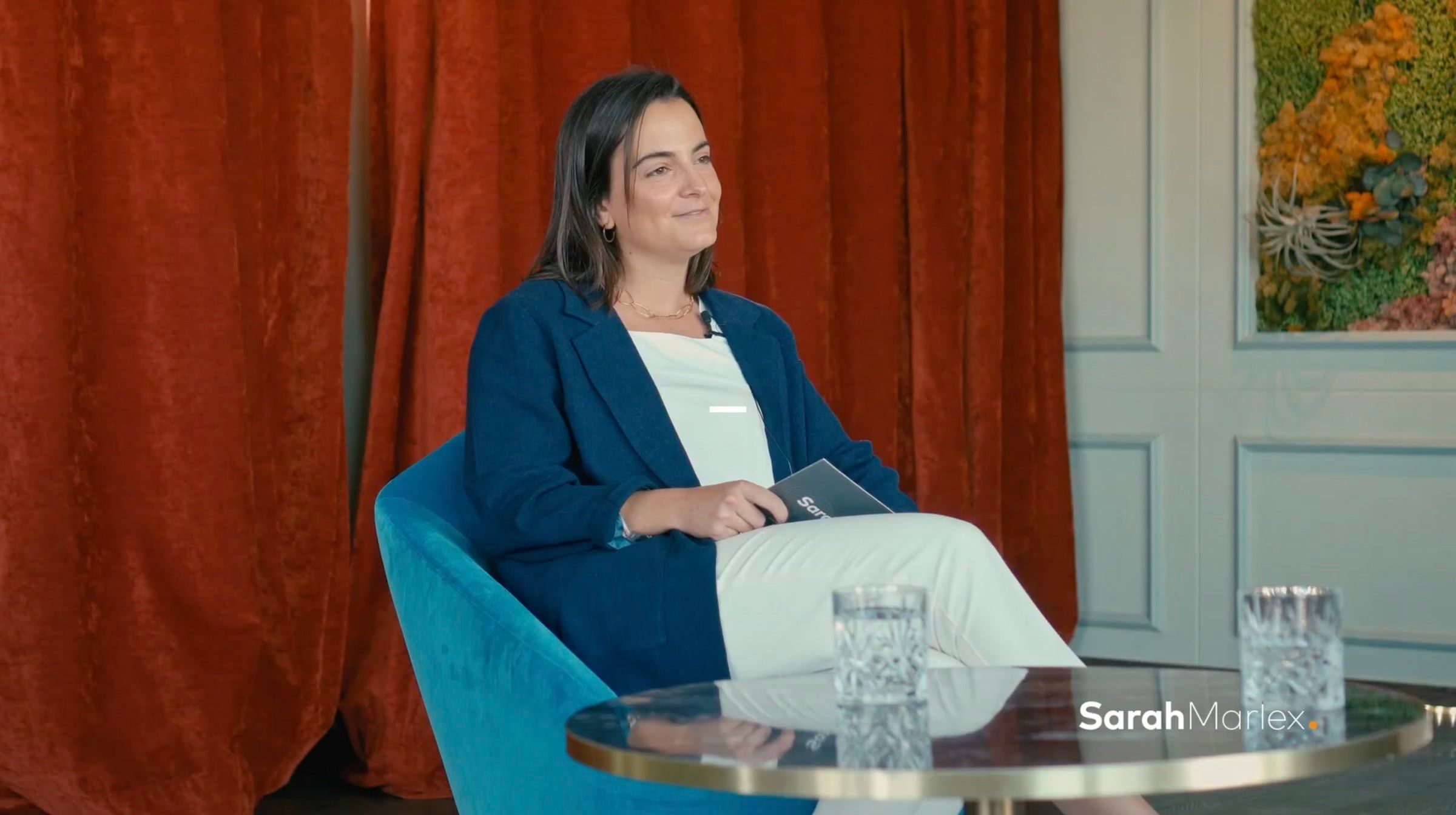 Woman in a navy blazer seated in a blue armchair in front of a red curtain, during the interview
